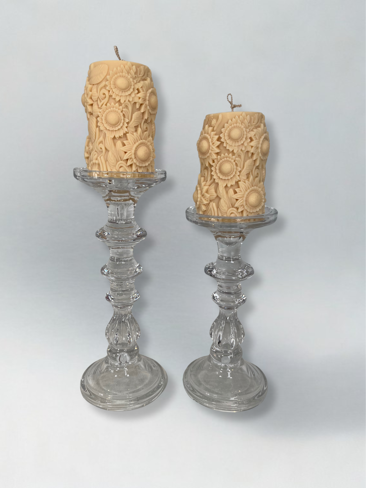 Two decorative candles embossed with sunflowers on glass candlesticks on a light gray background