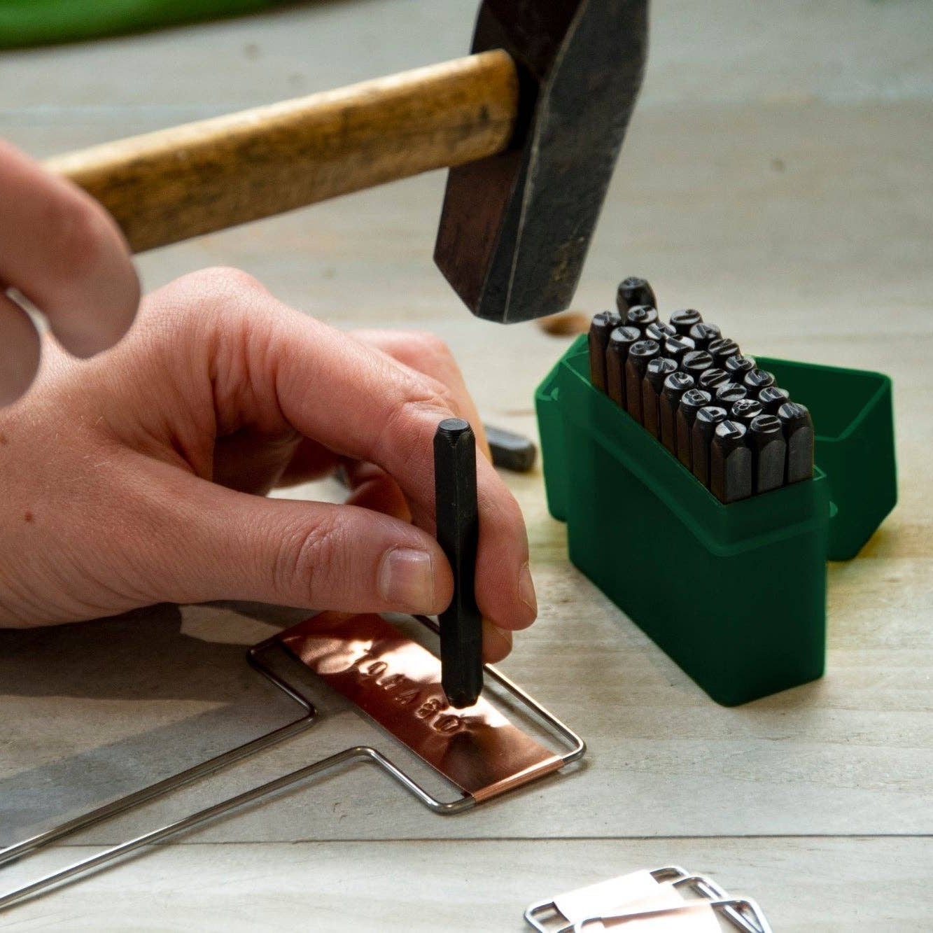 Person using a hammer and stamping tool on copper sheet with various tools on a wooden surface.