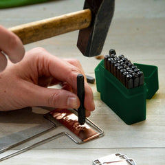 Person using a hammer and stamping tool on copper sheet with various tools on a wooden surface.