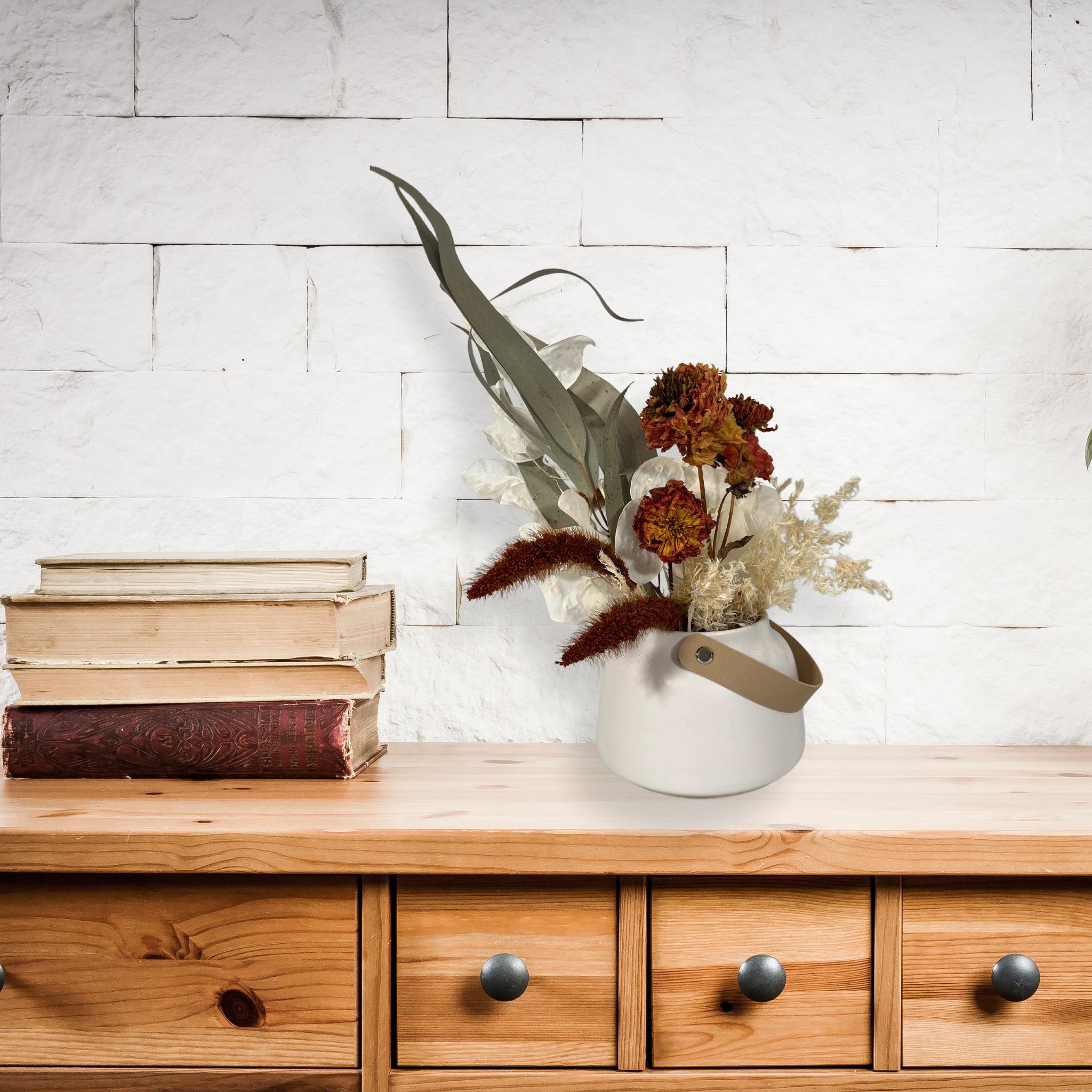 A floral arrangement on top of a wood console that has slender stems of natural green willow eucalyptus reach out from one side, complemented on the opposite side by the soft, bleached plumes of Calamgrostis canadensis. Tucked within the arrangement, ethereal Lunaria in pristine white adds a delicate, luminous quality. A cluster of five Dahlia stems, their petals a beautiful rust tone that compliment the demure matte white ceramic tapered vase.