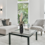 Modern living room with a glass coffee table, and patterned armchair. The focal point is a dried floral arrangement has soft, elegant plumes of natural Pampas Grass reach gracefully upward, complemented by the bold, preserved forms of blue Eucalyptus and the airy lightness of German Statice (sea lavender). The delicate whimsy of bleached Triticum (wheat) wisps adds a touch of rustic charm, while soft white Cape Flowers provide subtle pops of pure, natural colour. 