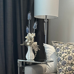 Living room corner with a vase of dried floral arrangement made with delicate, lace-like petals of white Cape Flowers against the intriguing, sculpted heads of blue Teasels. Adding airy lightness and enduring colour is German Statice. The room also has a lamp, and sofa with floral pillows.