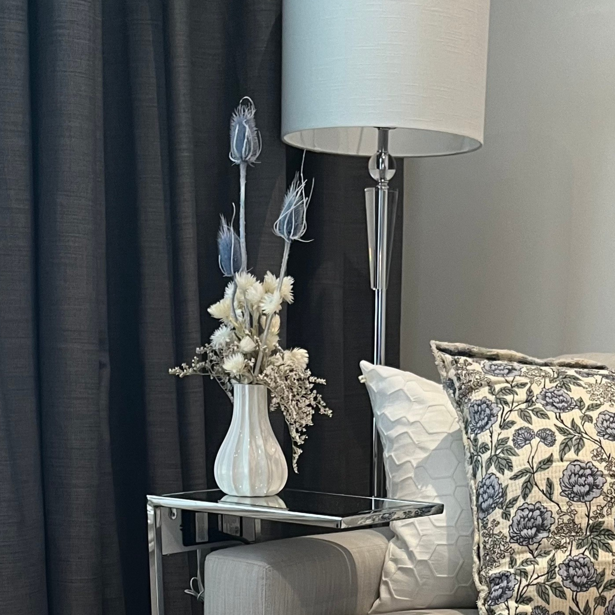 Living room corner with a vase of dried floral arrangement made with delicate, lace-like petals of white Cape Flowers against the intriguing, sculpted heads of blue Teasels. Adding airy lightness and enduring colour is German Statice. The room also has a lamp, and sofa with floral pillows.
