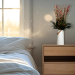 A bedroom scene acts as the background for a white ceramic vase with a leather handle acts a the vessel from which a beautiful wisp of blooms rises. The central feature is the delicate spires of natural pink Larkspur, reaching upwards with gentle movement. These are complemented by charming clusters of natural pink roses and a base of lush, preserved asparagus greens. Cheerful accents of sunny yellow Statice complete this delightful composition, capturing the essence of a blossoming garden.