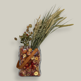 Decorative glass vase shaped like a paper bag with a leather handle filled with dried helichrysum, a beautifully arranged array of natural oats, Nigel, and safflower rise up from the vase.