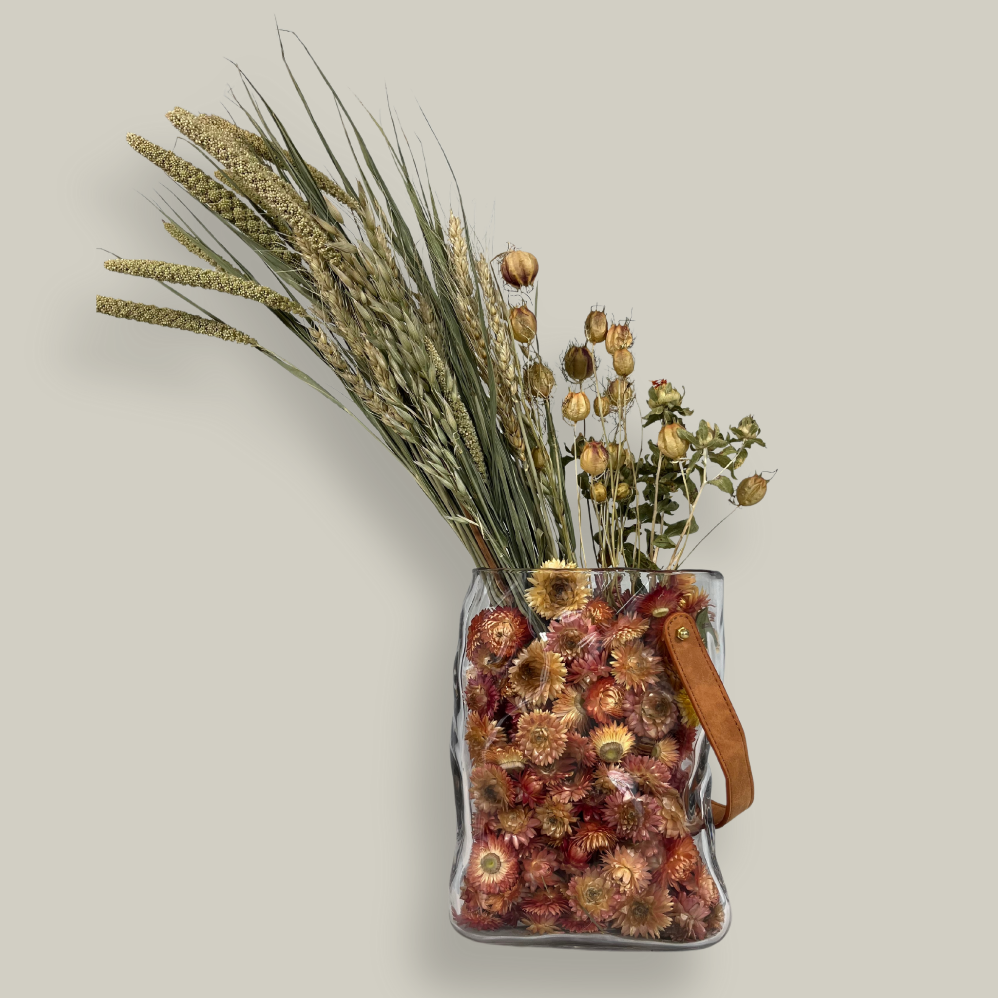 Decorative glass vase shaped like a paper bag with a leather handle filled with dried helichrysum, a beautifully arranged array of natural oats, Nigel, and safflower rise up from the vase.