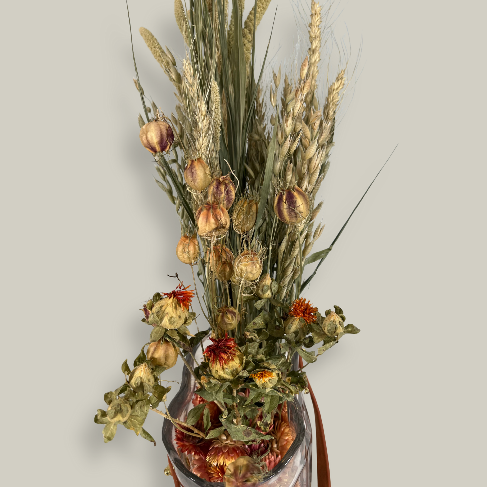 Decorative glass vase shaped like a paper bag with a leather handle filled with dried helichrysum, a beautifully arranged array of natural oats, Nigel, and safflower rise up from the vase.
