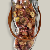 Decorative glass vase shaped like a paper bag with a leather handle filled with dried helichrysum, a beautifully arranged array of natural oats, Nigel, and safflower rise up from the vase.