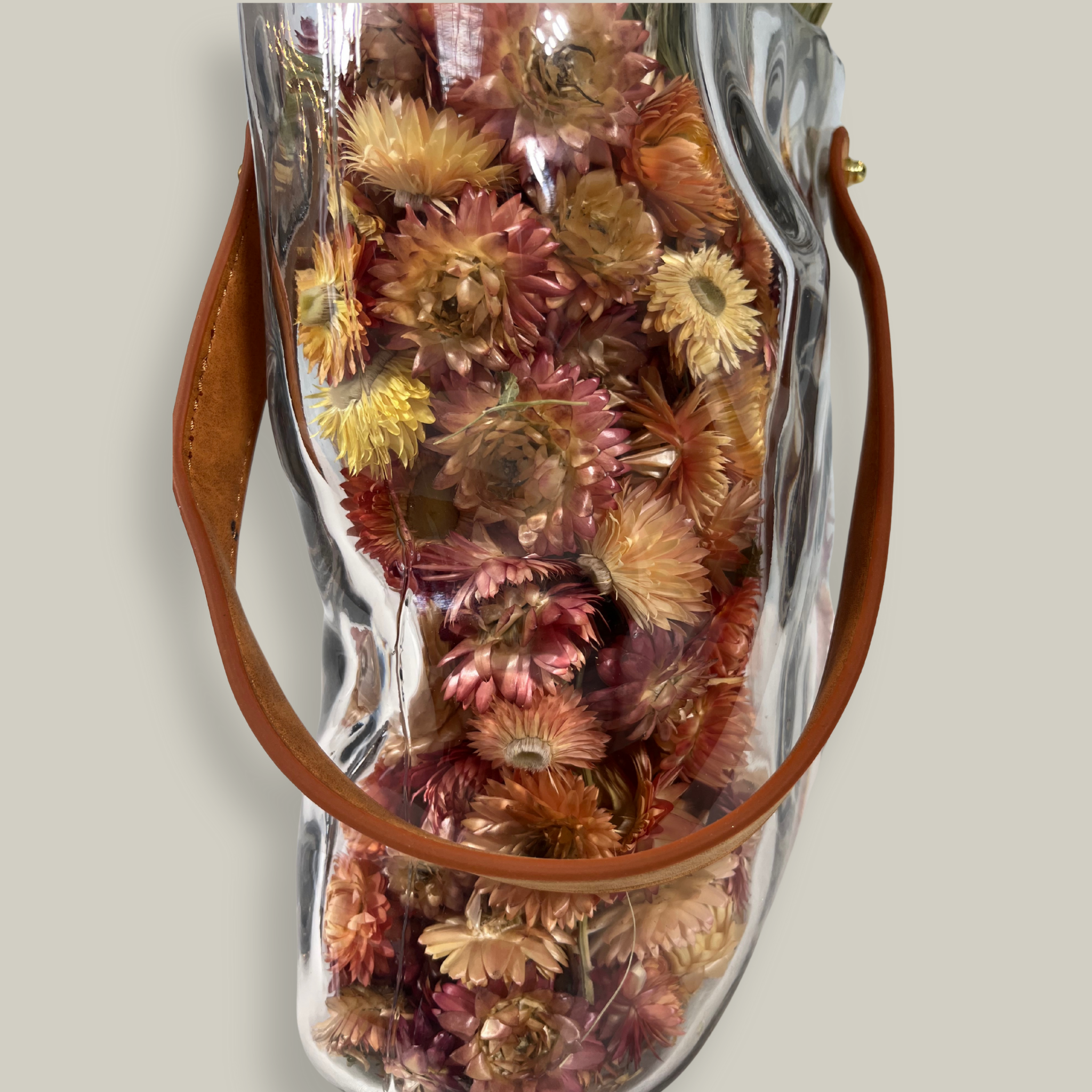 Decorative glass vase shaped like a paper bag with a leather handle filled with dried helichrysum, a beautifully arranged array of natural oats, Nigel, and safflower rise up from the vase.