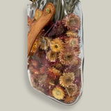 Decorative glass vase shaped like a paper bag with a leather handle filled with dried helichrysum, a beautifully arranged array of natural oats, Nigel, and safflower rise up from the vase.
