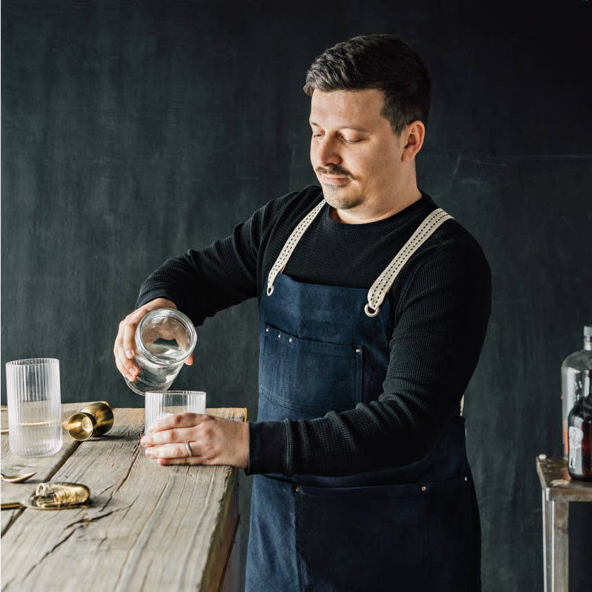 Man in a dark apron pouring a drink at a bar with bottles in the background