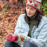 Woman wearing a beautiful hand knit floral hat, gloves, and denim jacket sitting on the ground with autumn leaves in the background.