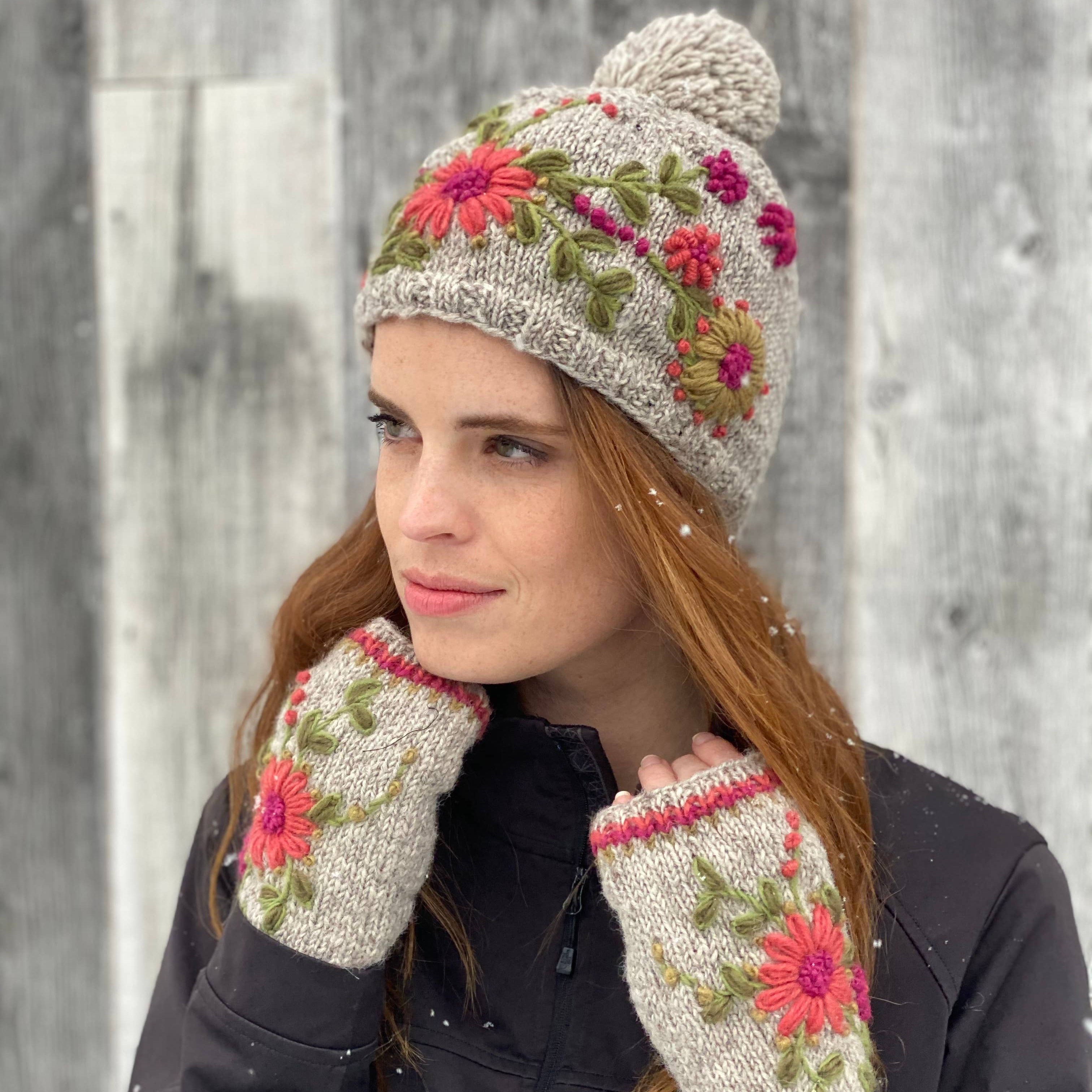 Woman wearing a floral knit hat with pom pom and hand=warmer style mittens against a snowy background