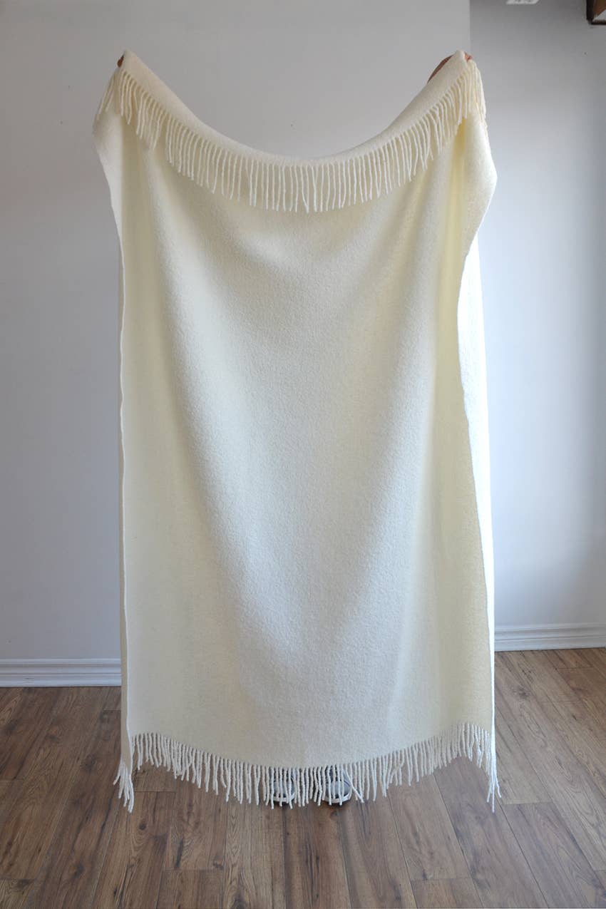 A luxurious wool blend boucle throw in ivory with fringes being held up by someone standing on a wooden floor against a white wall