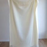 A luxurious wool blend boucle throw in ivory with fringes being held up by someone standing on a wooden floor against a white wall