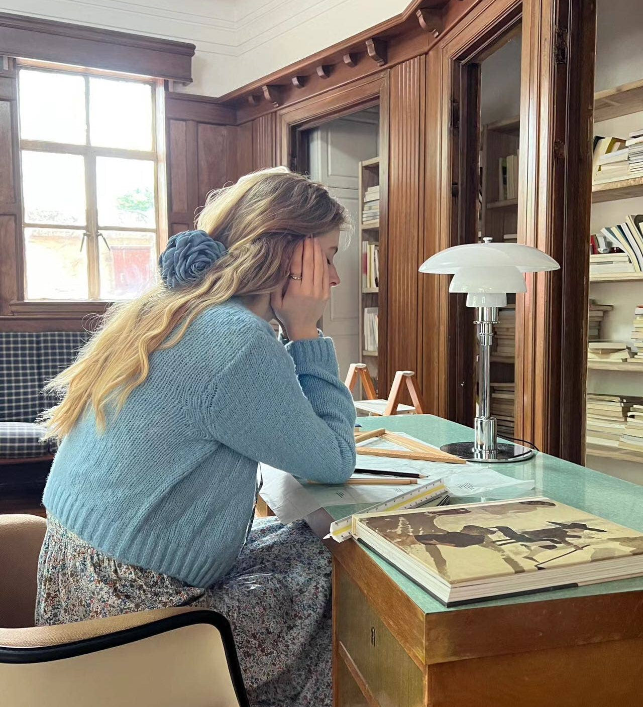 Woman wearing a blue floral hair clip sitting at a desk in a room with wooden paneling and books.