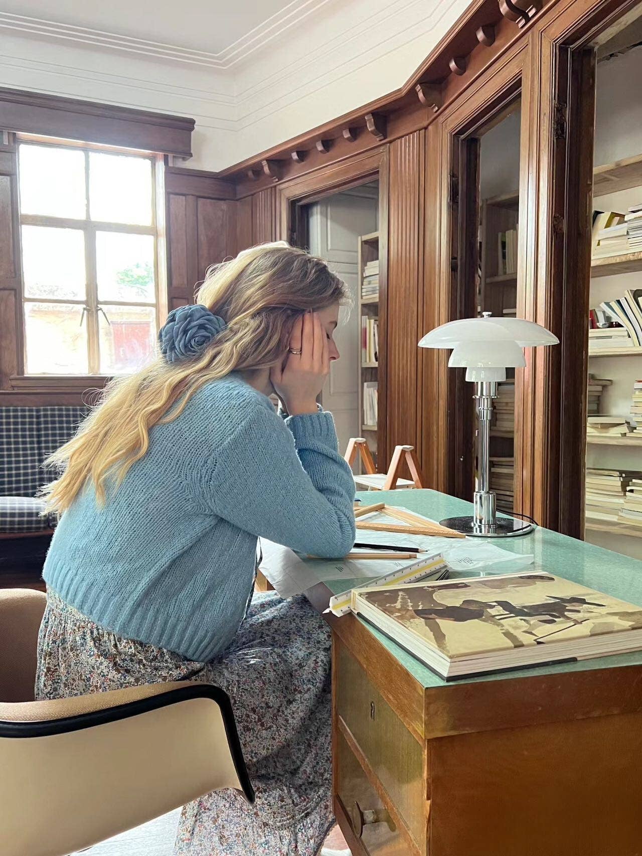 Woman wearing a blue floral hair clip sitting at a desk in a room with wooden paneling and books.