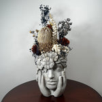A decorative sculptural head of a woman's face with hands holding it, made from light gray stone or resin, topped with dried flowers and botanicals including a large gold-toned protea and cream and rust blooms, displayed on a dark round table against a plain light wall.