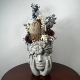 A decorative sculptural head of a woman's face with hands holding it, made from light gray stone or resin, topped with dried flowers and botanicals including a large gold-toned protea and cream and rust blooms, displayed on a dark round table against a plain light wall.