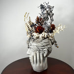 A sculptural dried flower arrangement in a pale gray stone-like head sculpture, displayed on a dark round table against a light gray wall.