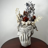 A sculptural dried flower arrangement in a pale gray stone-like head sculpture, displayed on a dark round table against a light gray wall.
