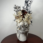 A decorative gray stone bust vase filled with dried floral arrangements in cream, gray, and dark brown tones, displayed on a round dark wood table against a plain light gray wall.