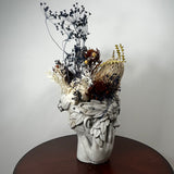 A sculptural ceramic head vase filled with dried botanicals, displayed on a round dark wood table against a plain light gray wall.