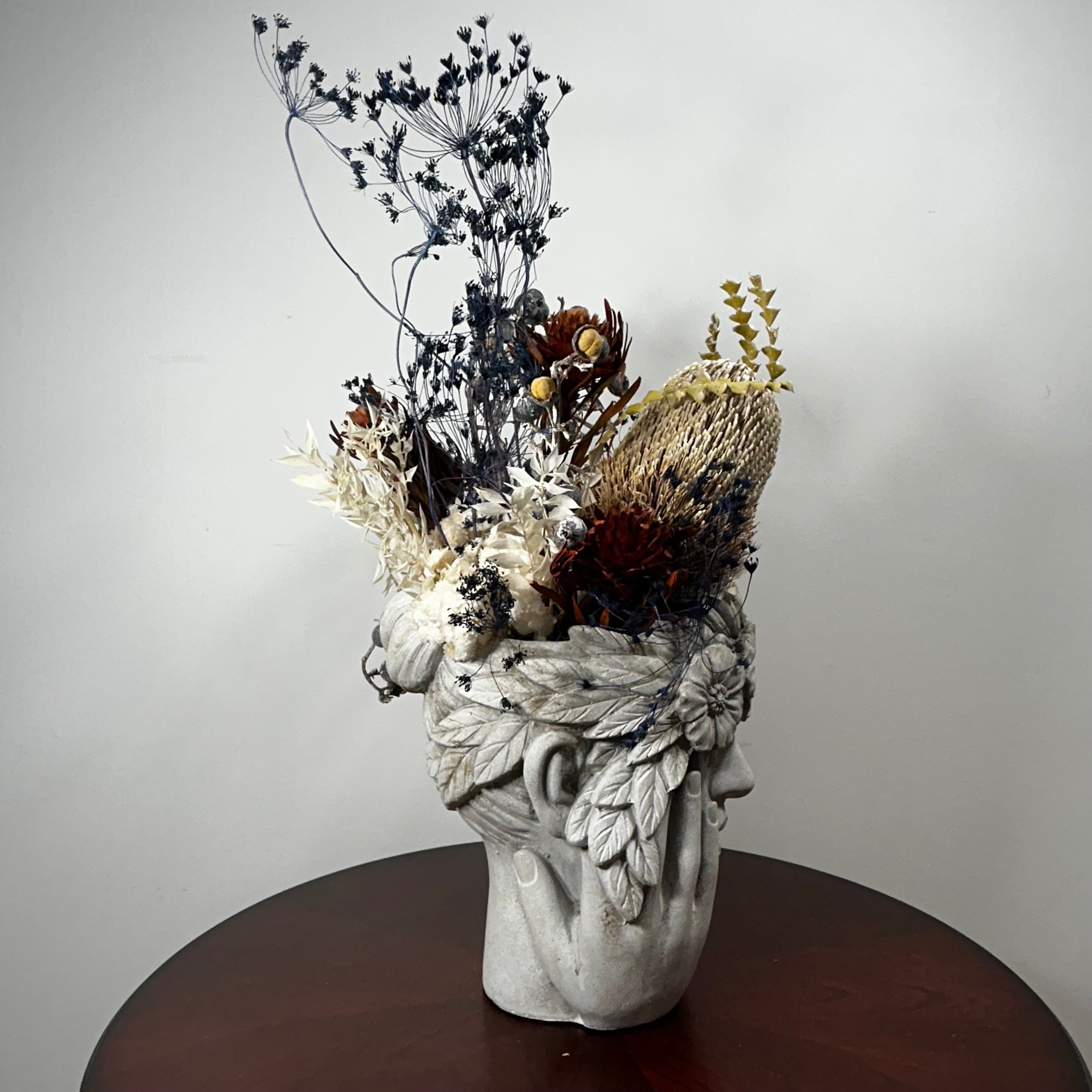 A sculptural ceramic head vase filled with dried botanicals, displayed on a round dark wood table against a plain light gray wall.