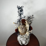 A decorative dried floral arrangement in a sculptural white flower-head vase, displayed on a round dark wooden table against a plain light gray wall.