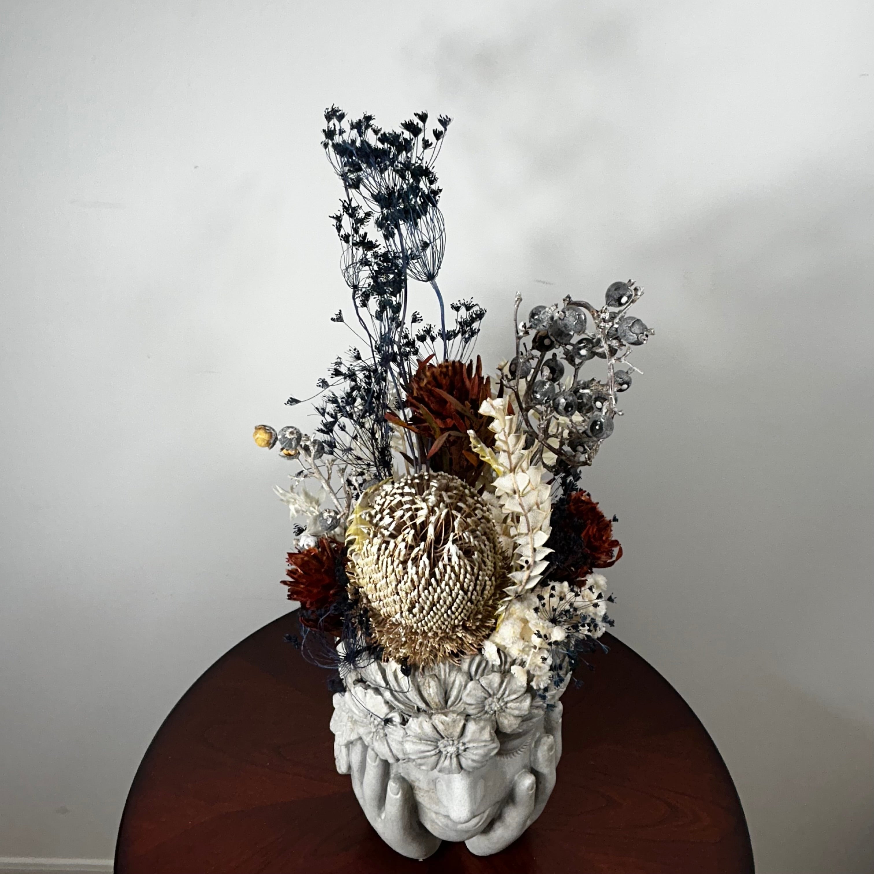 A decorative dried floral arrangement in a sculptural white flower-head vase, displayed on a round dark wooden table against a plain light gray wall.