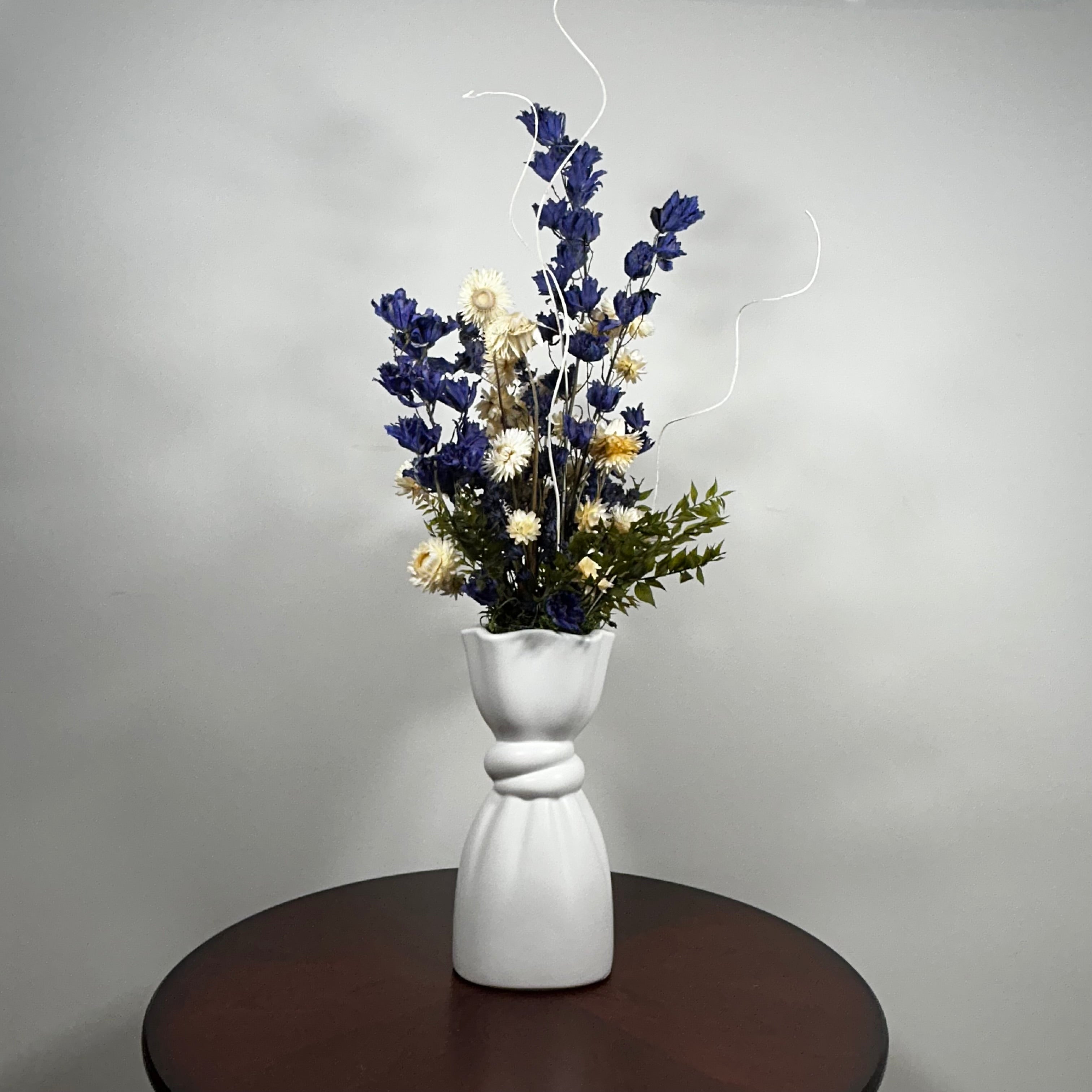 A white ceramic vase with a sculptural, twisted neck design, filled with a dried floral arrangement featuring dark blue, cream, and green elements, set on a dark brown round table against a neutral gray background.