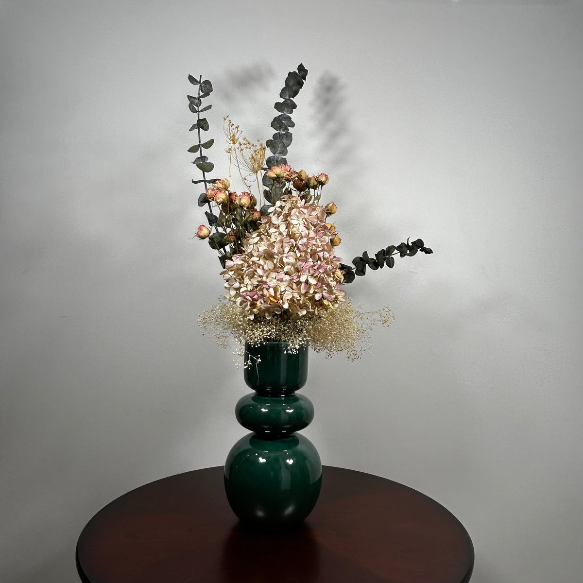 A green ceramic vase, filled with a dried floral arrangement featuring dark blue eucalyptus, dried hydrangea, and dried pink spray roses, and a cloud of preserved baby's breath, set on a dark brown round table against a neutral grey background.