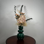 A green ceramic vase, filled with a dried floral arrangement featuring dark blue eucalyptus, dried hydrangea, and dried pink spray roses, and a cloud of preserved baby's breath, set on a dark brown round table against a neutral grey background. 1