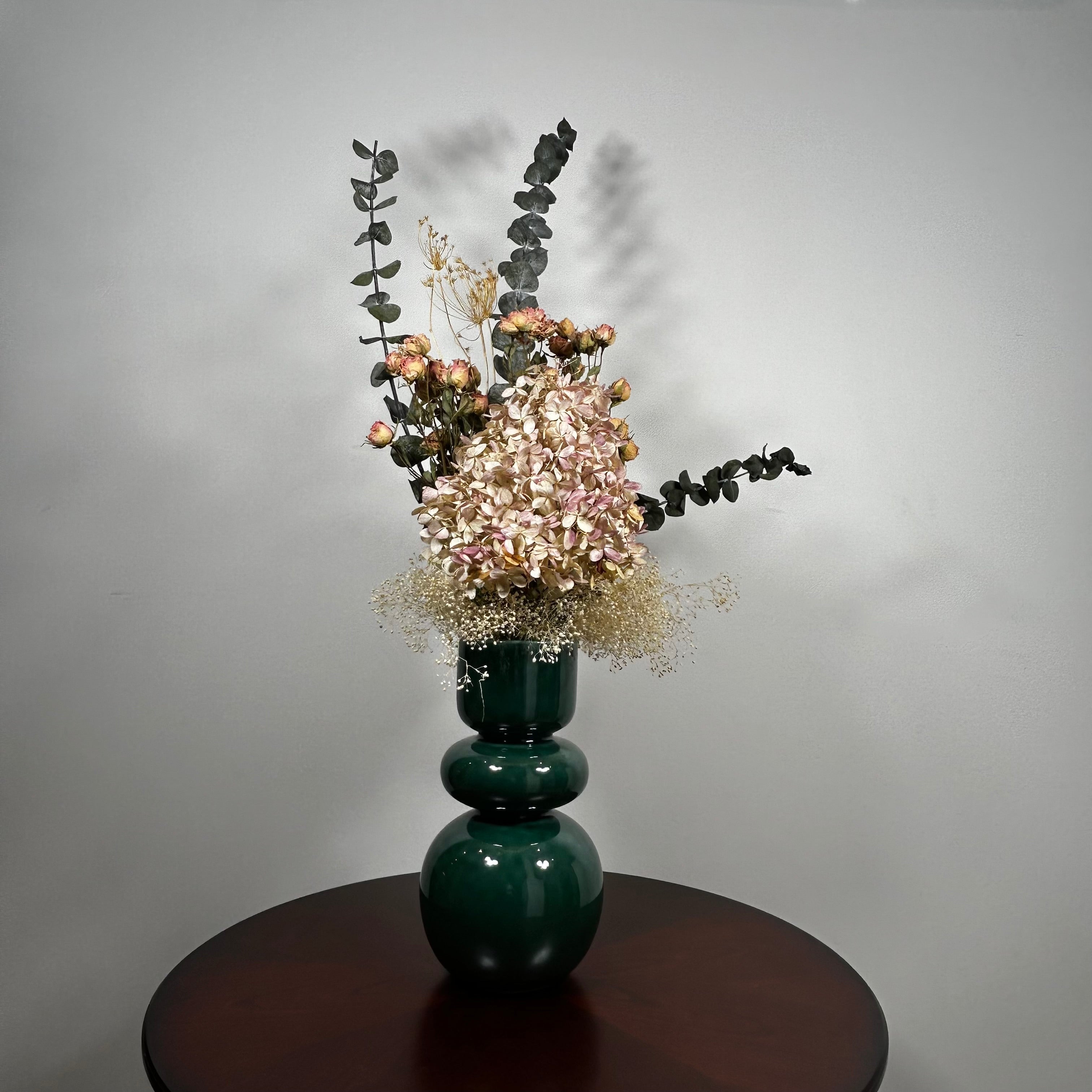 A green ceramic vase, filled with a dried floral arrangement featuring dark blue eucalyptus, dried hydrangea, and dried pink spray roses, and a cloud of preserved baby's breath, set on a dark brown round table against a neutral grey background.