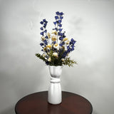 A white ceramic vase with a sculptural, face-like neck and flared rim, holding a dried floral arrangement of blue delphiniums, cream daisies, and green foliage, set on a dark round wooden table against a light gray wall.
