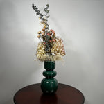 A green ceramic vase, filled with a dried floral arrangement featuring dark blue eucalyptus, dried hydrangea, and dried pink spray roses, and a cloud of preserved baby's breath, set on a dark brown round table against a neutral grey background. 4