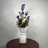 A white ceramic vase with a sculptural, flared top and ribbed base, holding a floral arrangement of blue delphiniums, cream daisies, and green foliage, set on a dark round wooden table against a light gray wall.