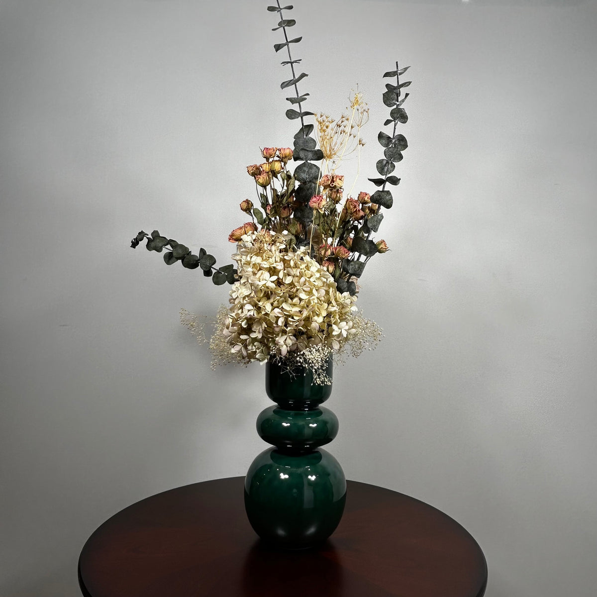 A green ceramic vase, filled with a dried floral arrangement featuring dark blue eucalyptus, dried hydrangea, and dried pink spray roses, and a cloud of preserved baby's breath, set on a dark brown round table against a neutral grey background.