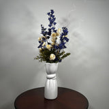 A white ceramic vase with a sculptural, flared base and a twisted center detail, filled with a dried floral arrangement featuring dark blue, cream, and green elements, set on a dark round wooden table against a neutral gray wall.