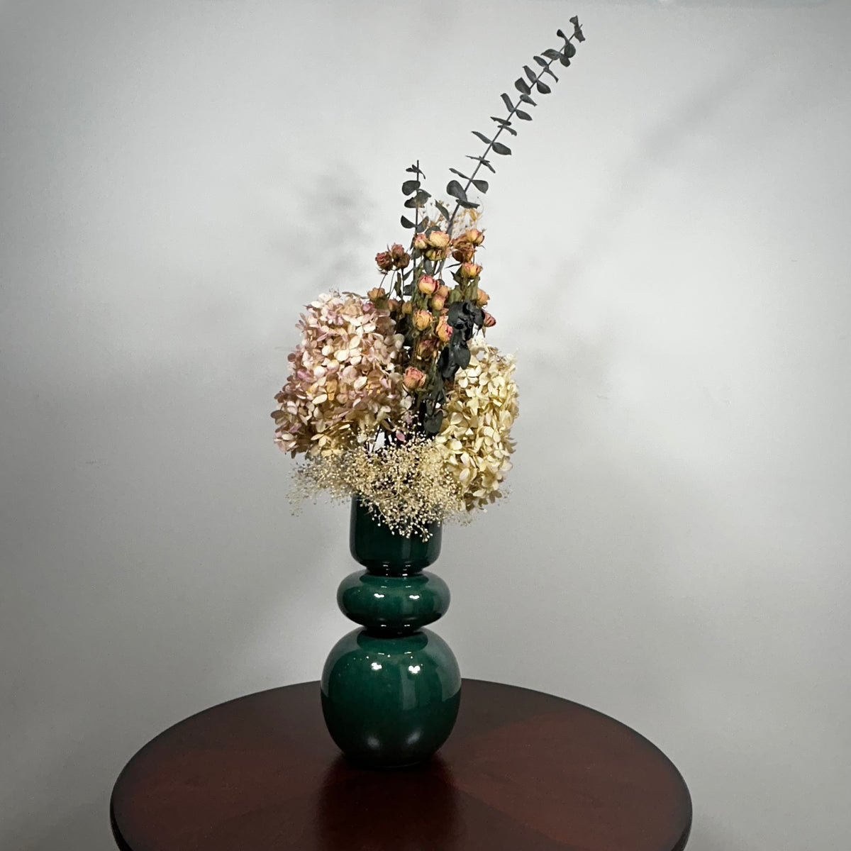 A green ceramic vase, filled with a dried floral arrangement featuring dark blue eucalyptus, dried hydrangea, and dried pink spray roses, and a cloud of preserved baby's breath, set on a dark brown round table against a neutral grey background.
