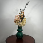 A green ceramic vase, filled with a dried floral arrangement featuring dark blue eucalyptus, dried hydrangea, and dried pink spray roses, and a cloud of preserved baby's breath, set on a dark brown round table against a neutral grey background. 2