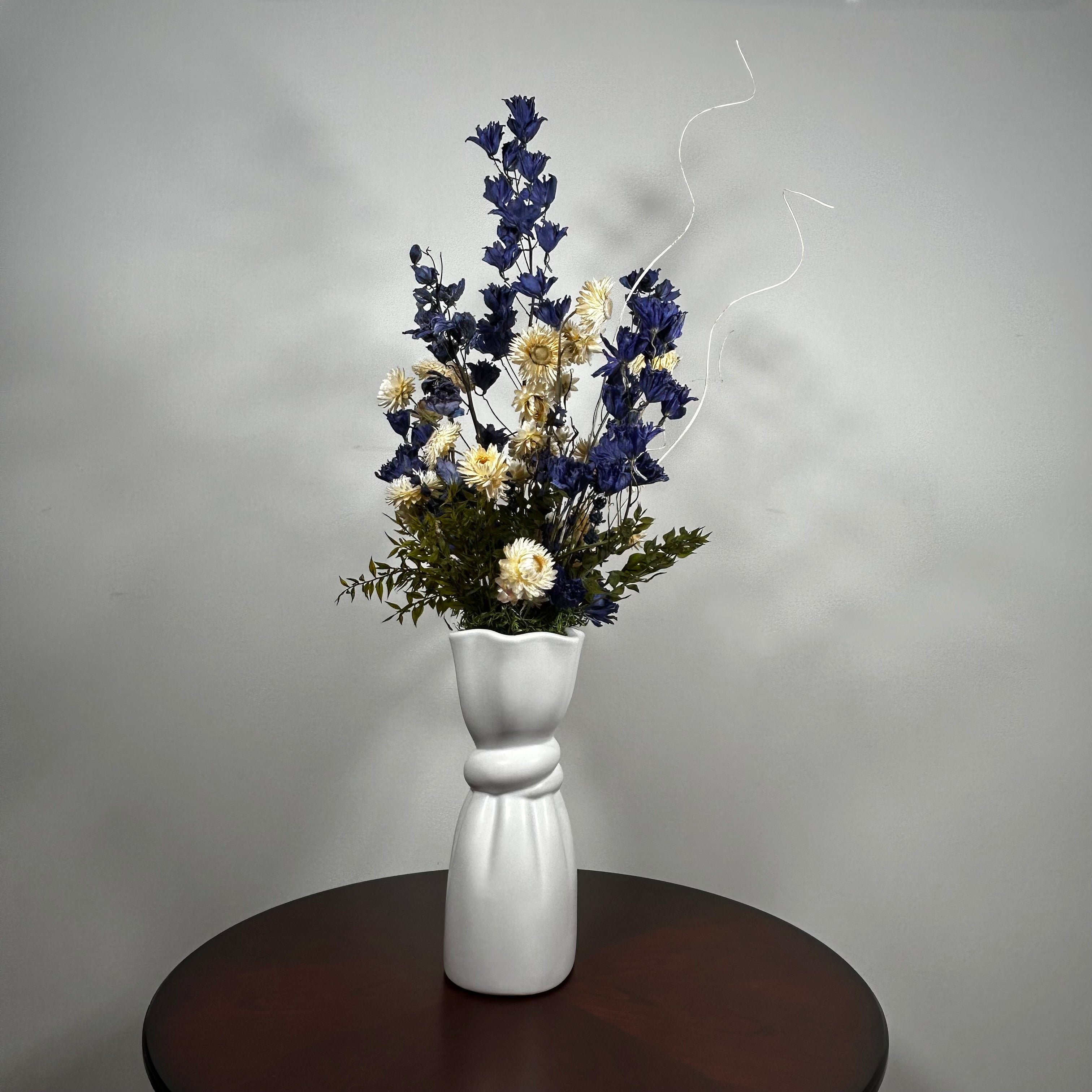 A white ceramic vase with a sculptural, flared base and a twisted center detail, filled with a dried floral arrangement featuring dark blue, cream, and green elements, set on a dark round wooden table against a neutral gray wall.