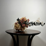 A dried floral arrangement with beige hydrangea-like blooms, burgundy roses, and green-gray eucalyptus stems, displayed on a dark round table against a plain light gray wall.