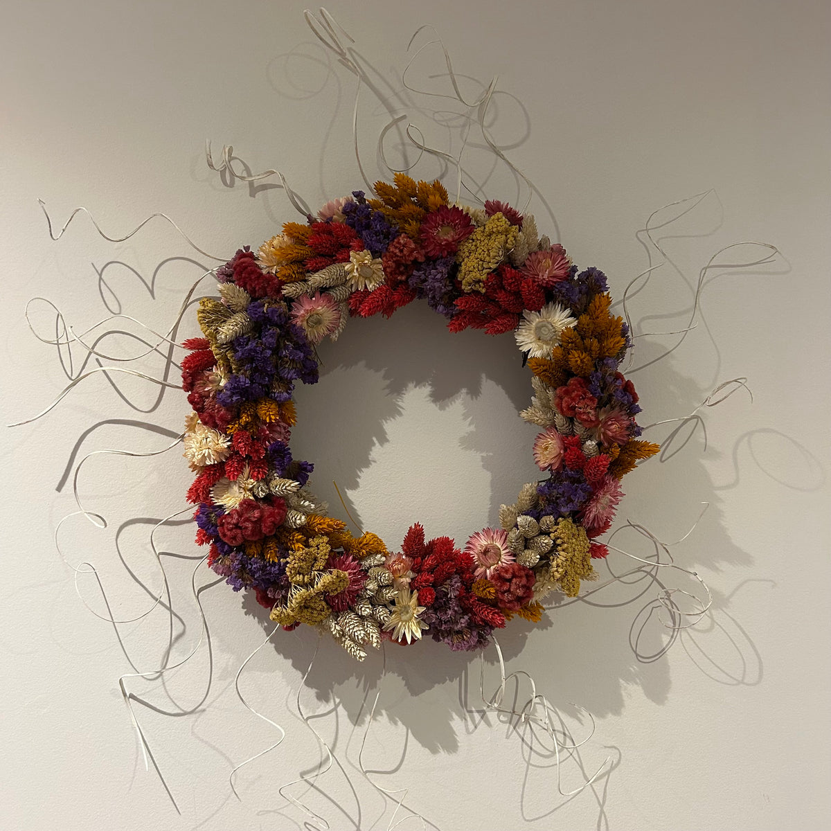 A circular floral wall art piece made of dried multicolored flowers and wrapped with thin silver wire, mounted on a plain light wall.