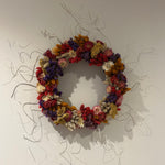 A circular floral wall art piece made of dried multicolored flowers and wrapped with thin silver wire, mounted on a plain light wall. 1