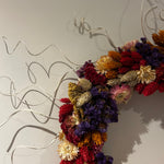 A dried floral wall sculpture featuring a dense cluster of multicolored artificial flowers and seed pods, accented by flowing white wire branches, mounted against a plain light gray wall. 2