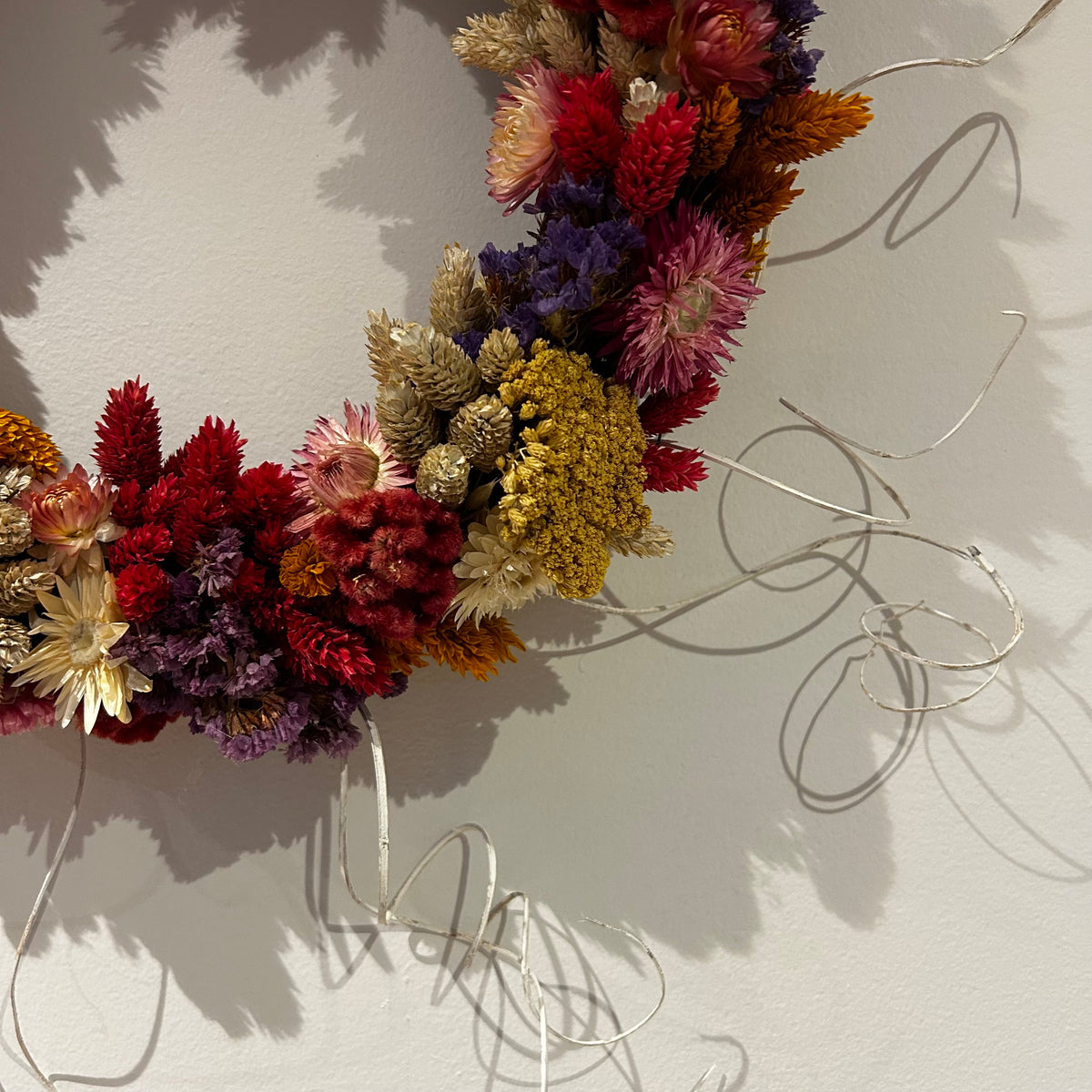 A dried flower wreath made from multicolored botanicals including red, purple, yellow, and beige blooms, attached to a thin silver wire frame and hung against a light wall.