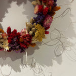 A dried flower wreath made from multicolored botanicals including red, purple, yellow, and beige blooms, attached to a thin silver wire frame and hung against a light wall. 3