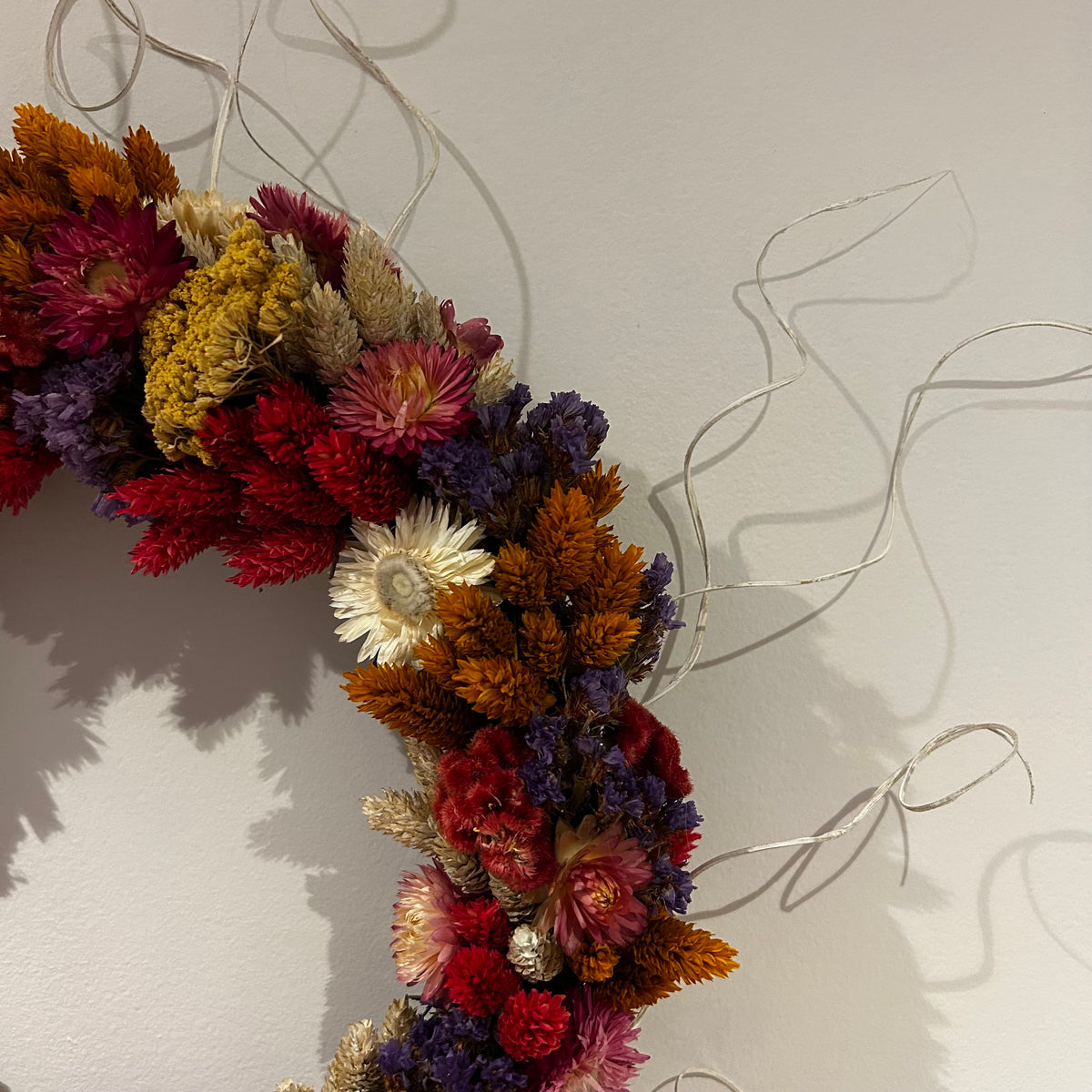 A dried flower wreath made of multicolored flowers including red, orange, purple, yellow, and white, hanging against a plain light gray wall with thin silver wire strands extending outward.