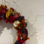 A dried flower wreath made of multicolored flowers including red, orange, purple, yellow, and white, hanging against a plain light gray wall with thin silver wire strands extending outward. 4